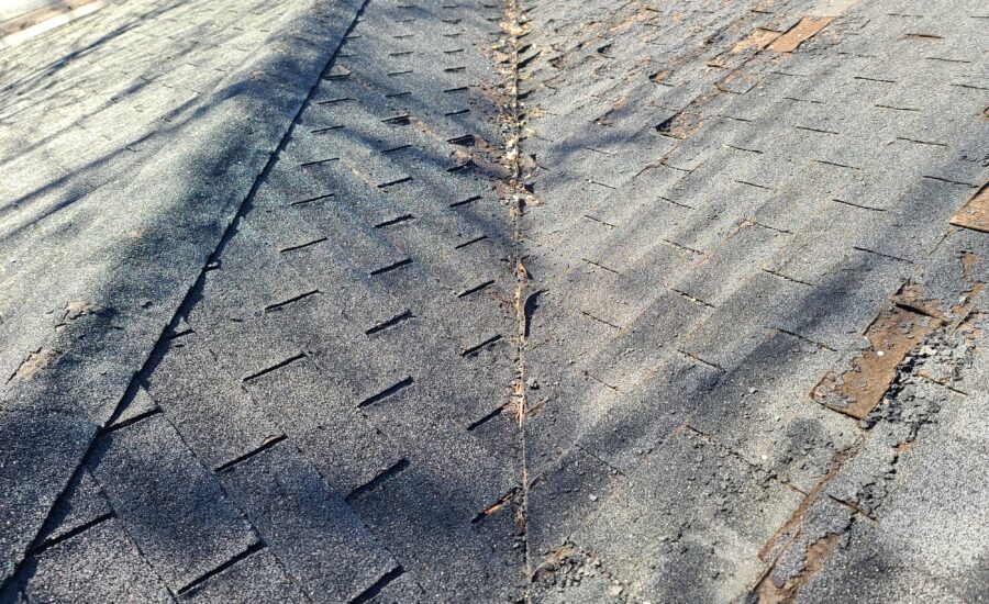 Close-up of weathered asphalt roof shingles with a narrow seam running down the center and small stones nearby.