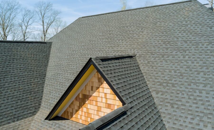 Steep gray shingled roof with a triangular wooden dormer exposing peach-colored wood shingles against a blue sky